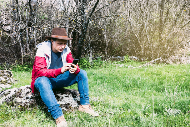 Entrepreneurial self-employed man wearing hat, looking at his mobile while relaxing in the countryside, sitting on a stone. Concept of work, enjoy, relax, Internet, technology and connectivity. - Foto, Imagem