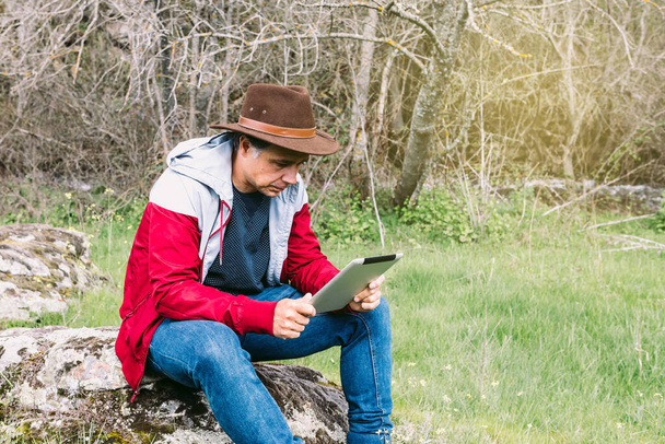 Empresario autónomo con sombrero, mirando su tableta mientras se relaja en el campo, sentado en una piedra. Concepto de trabajo, disfrute, relax, Internet, tecnología y conectividad. - Foto, imagen