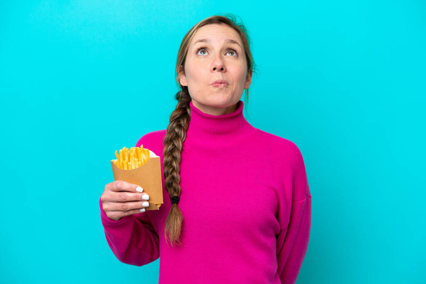 Young caucasian woman holding fried chips isolated on blue background and looking up - Fotoğraf, Görsel