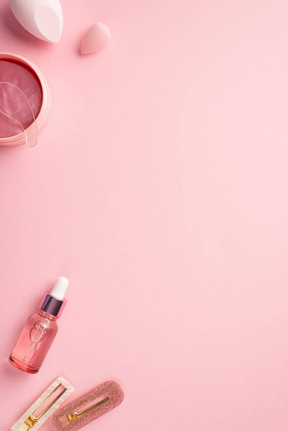 Beauty concept. Top view vertical photo of beauty blenders pink eye patches with special spoon pink transparent dropper bottle and two stylish hairpins on pastel pink background with empty space - Foto, Imagem
