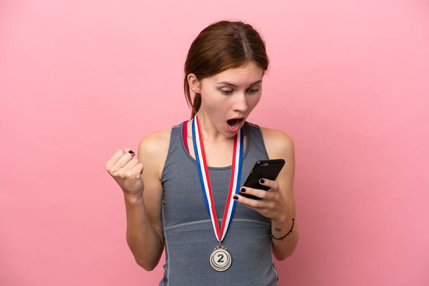 Young English woman with medals isolated on pink background surprised and sending a message - 写真・画像