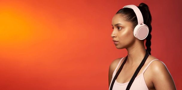 Playing tracks that keep her focused. Studio shot of a sporty young woman wearing headphones and posing with a skipping rope around her neck against a red background. - Foto, afbeelding