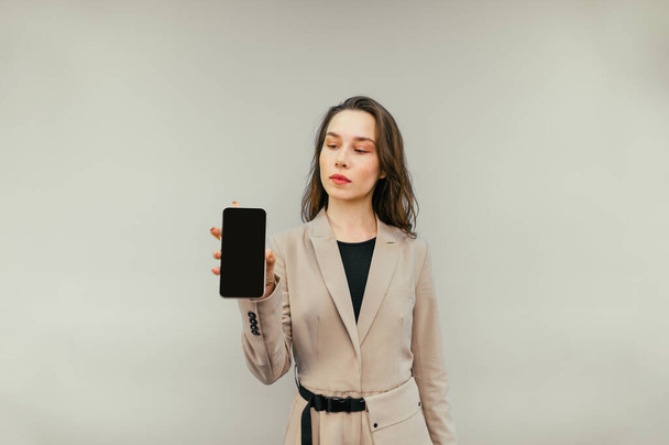 Красивая женщина в куртке стоит на бежевом фоне и показывает смартфон с черным экраном. - Фото, изображение