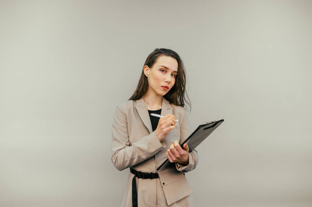 Serieuze zakenvrouw in een jasje staat met een papieren tablet en een pen in zijn handen en kijkt naar de camera op een beige achtergrond. - Foto, afbeelding