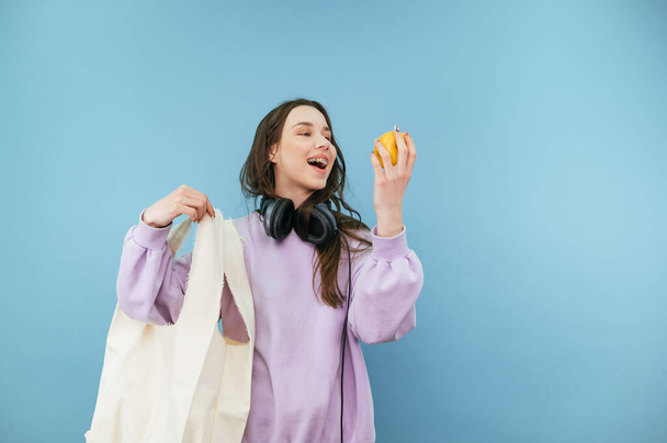 Atrakcyjna kobieta z eko-torbą w rękach stoi na niebieskim tle z jabłkiem w dłoni i patrzy na owoce z uśmiechem na twarzy. - Zdjęcie, obraz