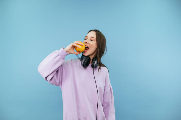 Beautiful woman in purple sweatshirt and headphones stands on a biting apple with a positive face on a blue background - Photo, Image