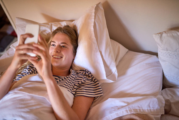 皆さんおはようございます。ベッドに横になりながら携帯電話を使って幸せな若い女性のショット. - 写真・画像