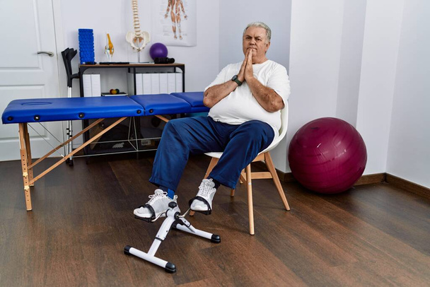 Senior caucasian man at physiotherapy clinic using pedal exerciser praying with hands together asking for forgiveness smiling confident.  - Photo, Image
