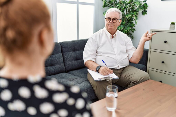Senior psychologist man at consultation office with a big smile on face, pointing with hand finger to the side looking at the camera.  - Foto, Imagem