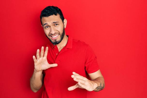 Latinomies parta yllään rento punainen t-paita inhottava ilme, tyytymätön ja pelokas tekee inhoa kasvot, koska vastenmielisyys reaktio. kädet ylhäällä  - Valokuva, kuva