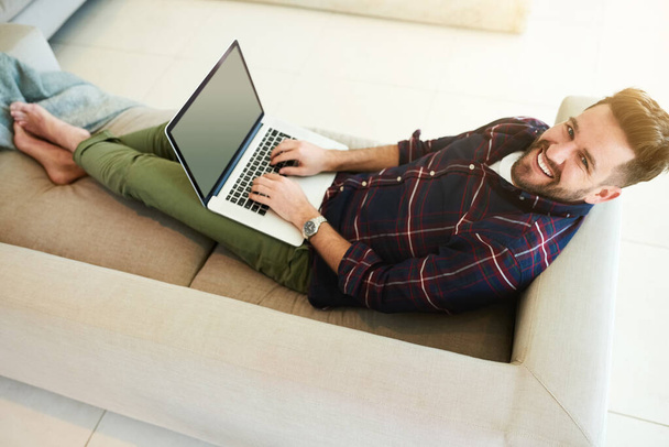En tarvitse muuta kuin laiskan päivän kotona. Laukaus rento nuori mies käyttää kannettavaa tietokonetta sohvalla kotona. - Valokuva, kuva