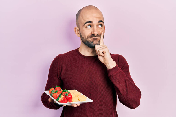 Молодий лисий чоловік їсть свіжі та здорові фрукти серйозне обличчя, думаючи про питання з рукою на підборідді, роздумуючи про заплутану ідею
  - Фото, зображення