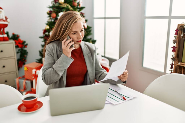 Joven mujer caucásica hablando en el teléfono inteligente trabajando sentado junto al árbol de Navidad en casa - Foto, Imagen