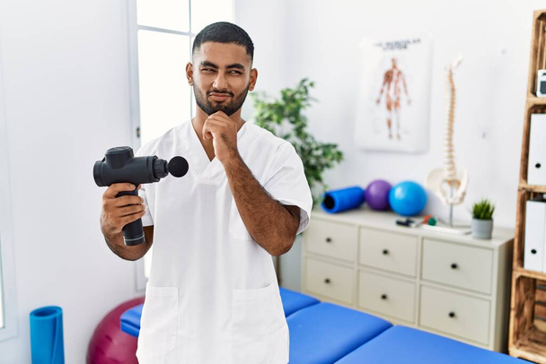 Young indian physiotherapist holding therapy massage gun at wellness center with hand on chin thinking about question, pensive expression. smiling and thoughtful face. doubt concept.  - Photo, Image