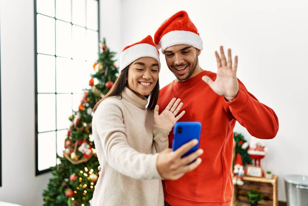 Молодая испаноязычная пара в рождественской шляпе с видеозвонком у смартфона дома. - Фото, изображение