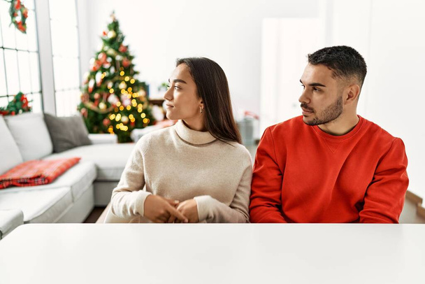 Jeune couple hispanique assis sur la table près de l'arbre de Noël regardant vers le côté, pose de profil relax avec visage naturel avec sourire confiant.  - Photo, image