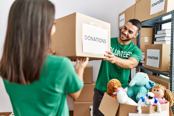 Молодая пара латинских волонтеров улыбается счастливо держа коробку пожертвований в благотворительном центре. - Фото, изображение