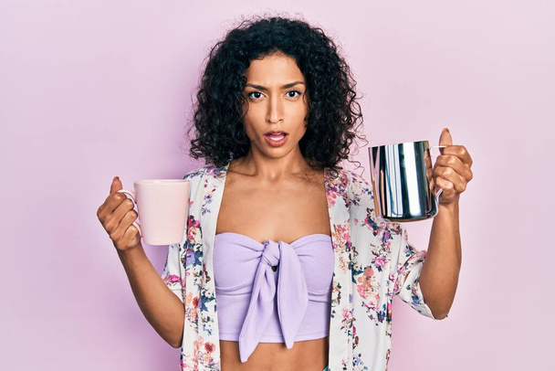 Jeune fille latine tenant une tasse de café et un pot de lait en état de choc, sceptique et sarcastique, surpris par la bouche ouverte  - Photo, image