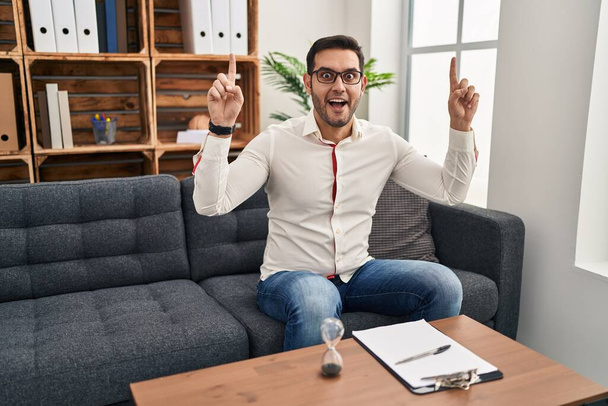Jonge Latijns-Amerikaanse man met baard werkt op consultatiebureau glimlachend verbaasd en verrast en wijzend met vingers en opgeheven armen.  - Foto, afbeelding