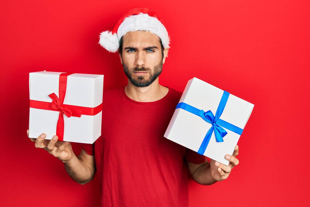 Junger hispanischer Mann mit Weihnachtsmütze und Geschenken in der Hand, skeptisch und nervös, die Stirn runzelnd wegen des Problems. negative person.  - Foto, Bild