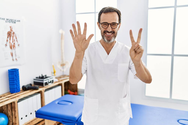 Middelbare leeftijd man met baard werken bij pijn herstel kliniek tonen en wijzen met de vingers nummer zeven, terwijl glimlachen vol vertrouwen en gelukkig.  - Foto, afbeelding