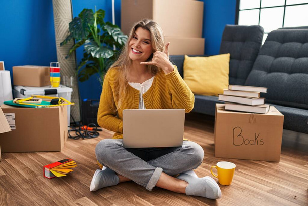 Young woman sitting on the floor at new home using laptop smiling doing phone gesture with hand and fingers like talking on the telephone. communicating concepts.  - 写真・画像