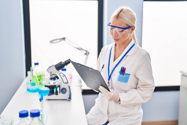 Giovane donna bionda con indosso uniforme da scienziato che scrive sugli appunti in laboratorio - Foto, immagini