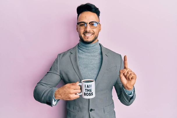 Joven árabe bebiendo de soy el jefe taza de café sonriendo con una idea o pregunta señalando el dedo con la cara feliz, número uno  - Foto, imagen