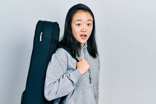 Fiatal kínai lány visel gitártok rémült és csodálkozott nyitott szájjal meglepetés, hitetlenség arc  - Fotó, kép