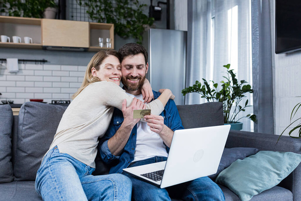 Online shopping. Man and woman shopping online from a notebook, holding a credit card in his hands. A purchase for a woman she chose herself. At home, sitting on the couch. Happy, satisfied - Foto, Imagen