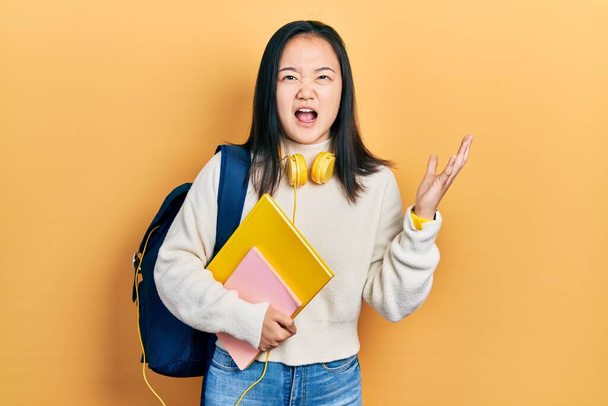 Młoda Chinka trzymająca studencki plecak i książki szalone i szalone krzyki i krzyki z agresywnym wyrazem twarzy i ramionami w górze. koncepcja frustracji.  - Zdjęcie, obraz