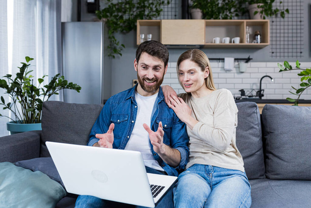 Young couple, family, man and woman sitting at home on the couch talking on video call from laptop to family, friends. They wave, smile, greet - Foto, Imagen
