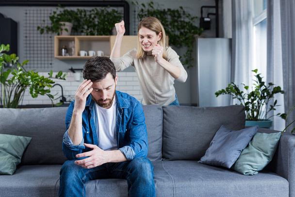 Young family, couple at home, man and woman quarrel, conflict, woman shouting and waving hands. Man sitting on sofa, holding head, listening - Foto, afbeelding