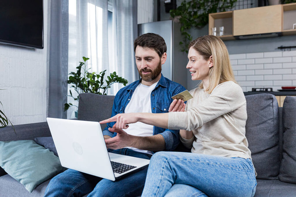 Online shopping. Man and woman shopping online from a notebook, holding a credit card in his hands. A purchase for a woman she chose herself. At home, sitting on the couch. Happy, satisfied - 写真・画像