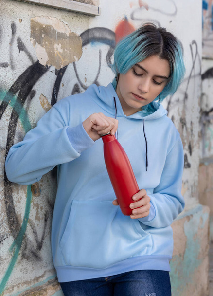 Teenager in blue hoodie opening red water bottle and looking down.Blue haired teen girl staying outdoors near graffiti wall. Clothing and reusable bottle mockup.  Hipster and adolescence concept - Foto, imagen