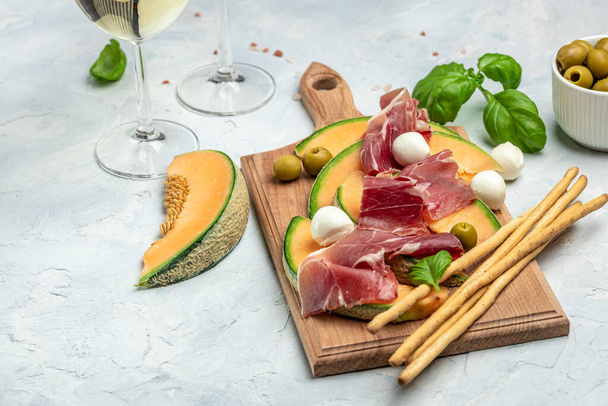 mediterranean salad with prosciutto or jamon, mozzarella and green basil leaves and Cantaloupe melon on white table, traditional Spanish and Italian appetizer served with wholemeal grissini, top view. - Photo, image