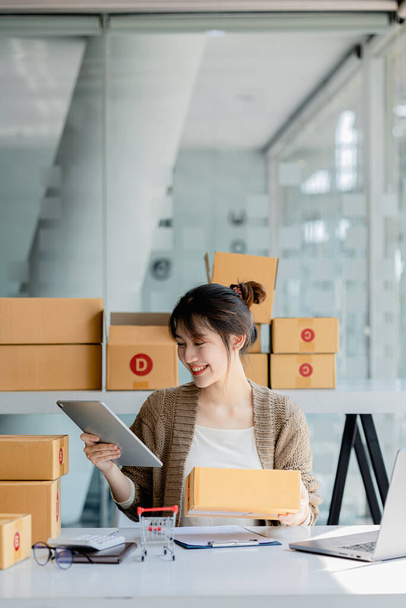 A beautiful Asian business owner opens an online store, she is checking orders from customers via tablet, sending goods through a courier company, concept of a woman opening an online business. - Photo, image