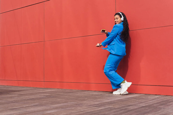 Full body side view of positive African American female in blue suit dancing while listening to music in headphones on red background - Foto, Bild