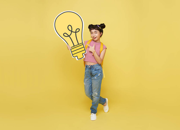 Щасливий азійський студент, що тримає лампочку зі шкільним мішком ізольованим на жовтому тлі. ідеї та творчий концепт. - Фото, зображення