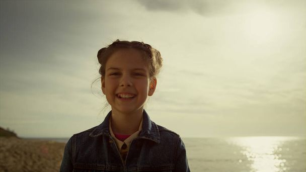 Szczęśliwa dziewczyna uśmiechnięta ciesząca się morską plażą rano. Dzieciak oglądający kamerę na brzegu. - Zdjęcie, obraz