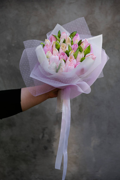 Ramo de malvaviscos. Tulipanes en un paquete festivo con una cinta blanca. La mano femenina sostiene flores. Sobre el fondo de una pared gris con una textura áspera.  - Foto, imagen