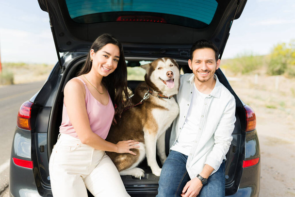 若い夫婦と犬が車のトランクの中で一緒に座っている間、道路旅行中に運転から休憩を取ります - 写真・画像