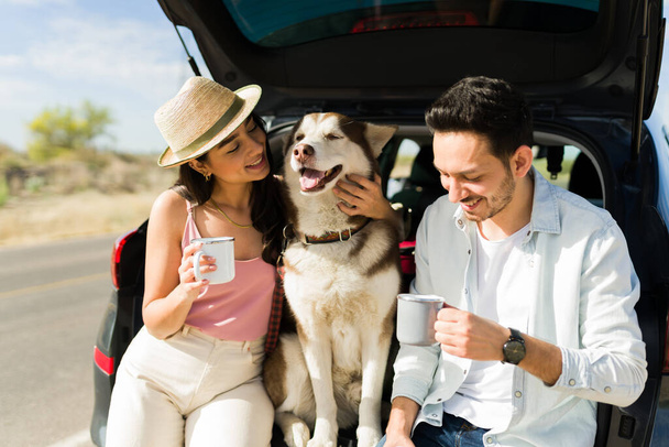 Χαρούμενο ζευγάρι χαϊδεύει το σκύλο τους και πίνει καφέ ενώ κάνει ένα διάλειμμα στο αυτοκίνητο κατά τη διάρκεια ενός ταξιδιού - Φωτογραφία, εικόνα