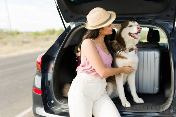 Dog lover. Gorgeous hispanic woman hugging her husky dog while sitting on the car trunk during a road trip - Fotó, kép
