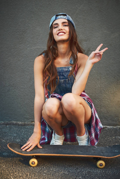 She has all the right moves. Portrait of a cheerful young woman seated on a skateboard while showing the peace sign outside during the day. - Foto, Imagem