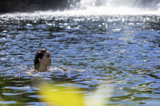A brunette woman refreshes herself by bathing in a river near a big waterfall of the cerrrado biome in west-central Brazil. Leisure. Lifestyle. Life in Nature.  Nature journey. - 写真・画像