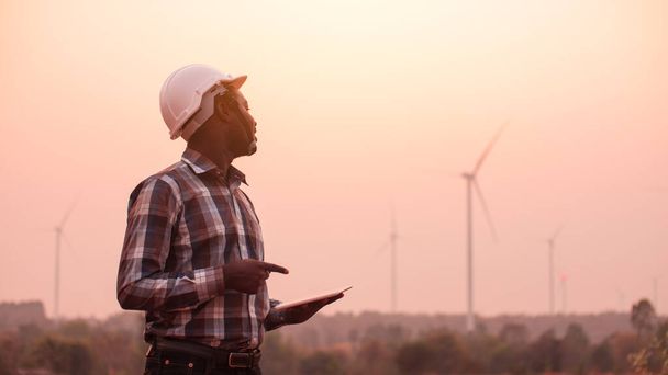Un ingénieur africain tient sa tablette devant les éoliennes générant une centrale électrique au coucher du soleil. - Photo, image
