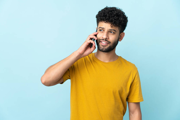 Joven hombre marroquí aislado sobre fondo azul manteniendo una conversación con el teléfono móvil con alguien - Foto, imagen