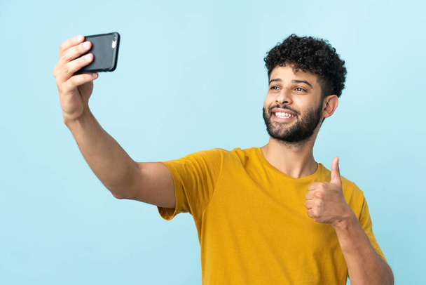 Молодой марокканский мужчина на синем фоне делает селфи с мобильным телефоном - Фото, изображение
