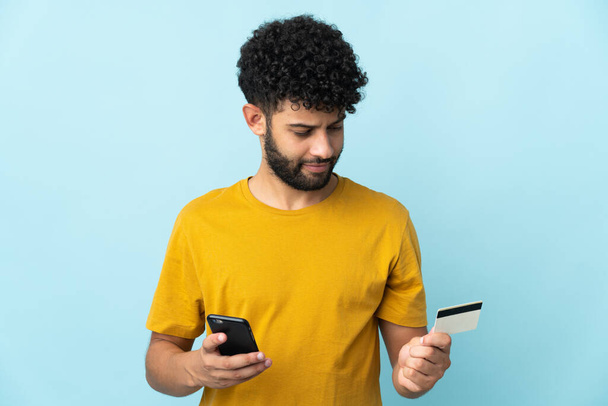 Young Moroccan man isolated on blue background buying with the mobile with a credit card - Foto, Imagem